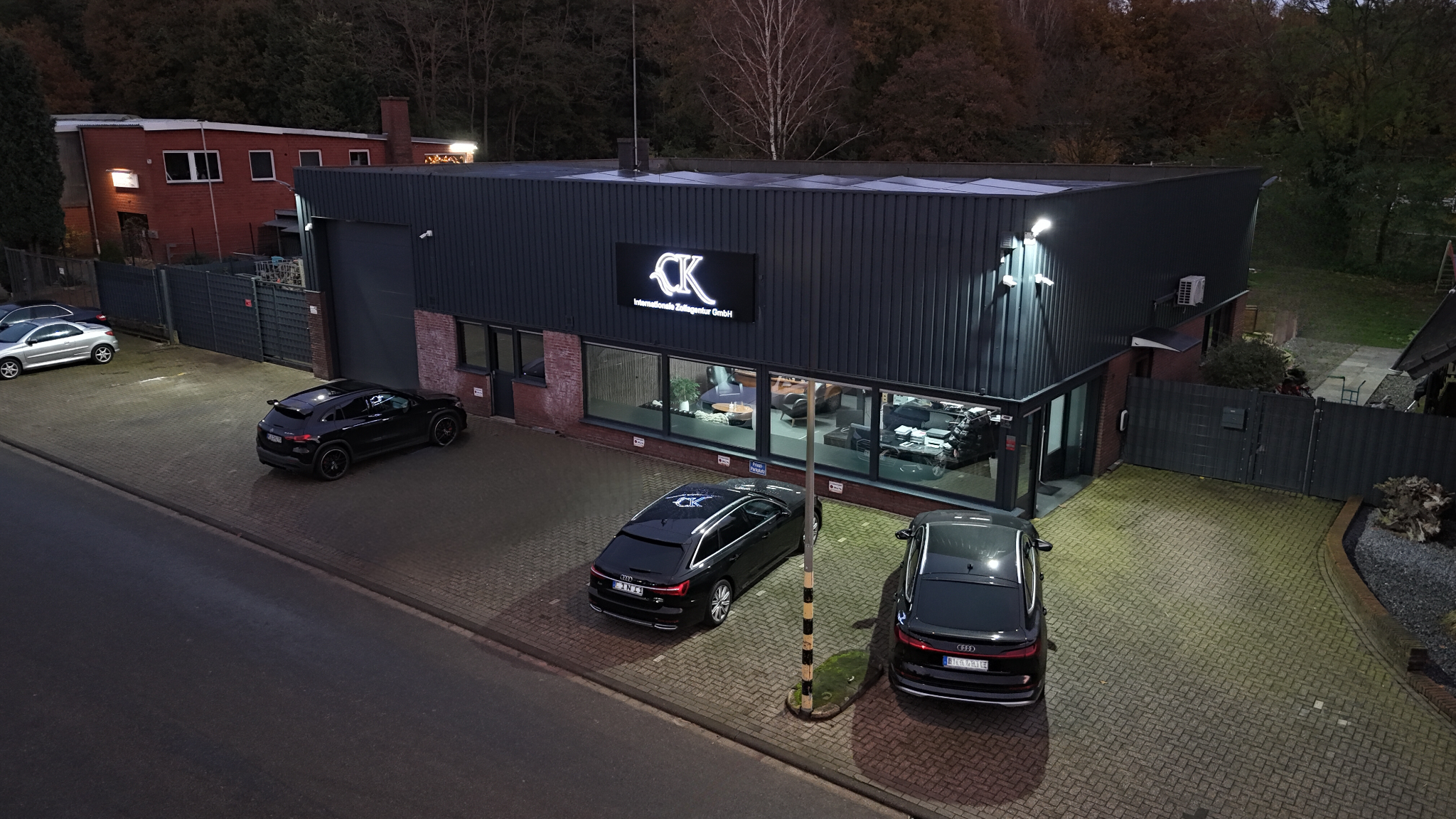 Das Bild zeigt ein Gebäude mit moderner Außenfassade und dem beleuchteten Firmenschriftzug "CK Internationale Zollagentur GmbH". Im Vordergrund sind mehrere geparkte Autos sichtbar, und die Umgebung weist auf einen Industrie- oder Gewerbepark hin. Das Gebäude hat große Fenster, die Einblicke in den Innenraum gewähren, in dem Tische und Büroausstattung zu erkennen sind. Es ist ein gut beleuchteter Parkplatz mit einer gepflegten Umgebung.